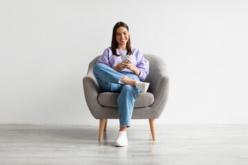 Young woman sitting in armchair, looking at mobile phone screen, using new cool application against...