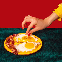 Closeup girl's hand tasting bacon and eggs isolated on green and red background. Vintage, retro style interior. Food pop art photography.