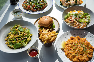 Gourmet foods on the table include burgers, pasta, french fries, vegetable salad, chicken salad, cauliflower, macaroni