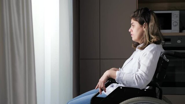 Young disabled woman with cerebral palsy sits in wheelchair near window at home.