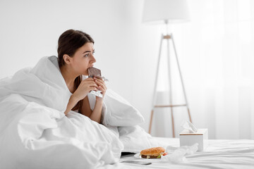 Disappointed upset millennial woman eating chocolate bar on bed with fast food at home, suffering...