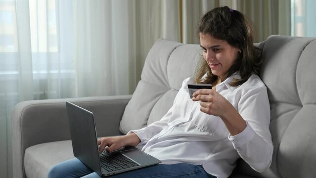 Young disabled woman with organic damage of central nervous system enters bank credit card details for salary payment on laptop sitting on sofa at home.