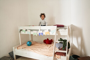 Multiracial boy looking at the camera with happy smile while standing at the second tier of his bed