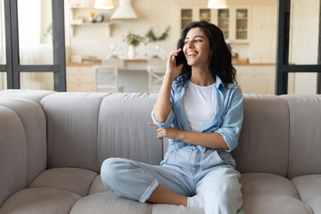 Fototapeta premium Happy young Caucasian lady sitting on couch, having mobile phone conversation at home, free space