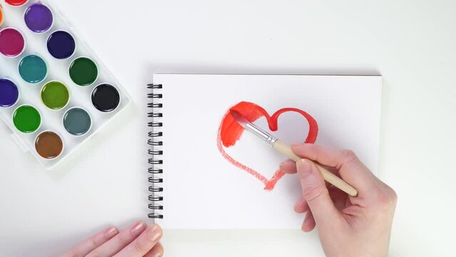 draw heart with paints and brush. paints and notebook for creativity. draw in sketchbook with watercolors. love is everywhere. hands paint red picture. caucasian female hand is engaged in creativity