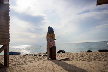 a young girl is sitting on a suitcase on a sandy seashore in a sundress and with a hat    