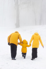 Father, mother and son in yellow jacket back, outdoor family time, woman, man holding hands with child. Snowy winter day