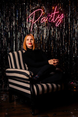 Elegant woman in black clothes sits in a chair. Party lettering on the wall. Red hair. Bright, shiny background.