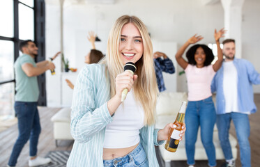 Carefree millennial blonde woman with bottle of beer singing karaoke on party with her interracial friends at home