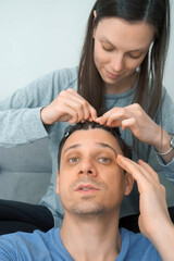 Man blogger is speaking at camera while woman is doing his hair french braids. He is smiling and talking recording his beauty vlog, vertical video. She is sitting on couch.