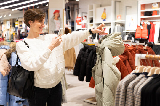 Photo of young attractive brunette woman with a short haircut in a white sweater chooses stylish and casual clothes in a store in a shopping center using mobile phone