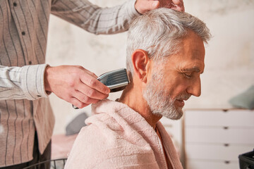 Elderly handsome man getting his hair and beard cut at home while his careful son helping