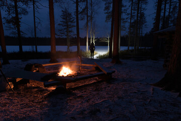 Figure standing on snowy path with headlamp near frozen lake and trees. Campfire and benches in the foreground