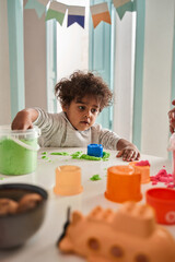 Brunette curly child playing with green kinetic sand and looking at something with interest