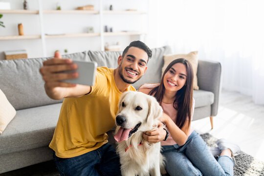Young affectionate multiracial couple taking selfie with pet dog at home