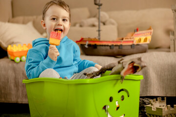 Little caucasian boy with blue hoodie playing with colourful toys and eating ice cream at home. Child having fun. Happy and cheerful kid plays with ship, dinosaurs, castle. Leisure activity.