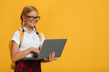 Cheerful smart caucasian young girl student in glasses studying with laptop, isolated on yellow background