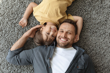 Happy dad and son. Top view of young positive father and his little son smiling at camera, lying on carpet floor - Powered by Adobe