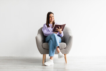 Weekend pastimes. Young Caucasian woman with open book sitting in comfy armchair against white...