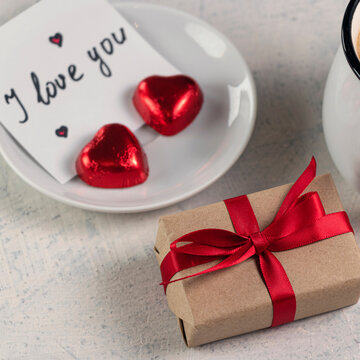 Square photo with gift with a red ribbon, a mug of coffee, heart-shaped chocolates and a note with the word I love you. Valentine's day breakfast. Valentine's Day concept. Soft focus