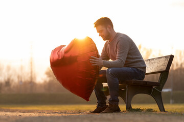 Sad young man holding up a heart-shaped balloon sitting on a bench in the park