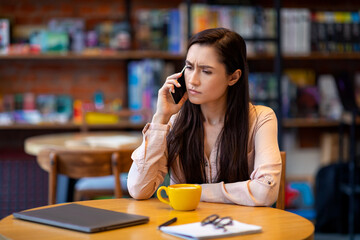 Angry freelancer arab lady talking to clients on cellphone while working remotely at cafe, using laptop computer