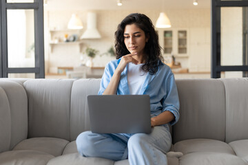 Serious young woman sitting on couch with laptop computer, working or studying from home