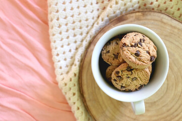 Mug filled with chocolate chip cookies and soft knitted blanket on a bed. Flat lay.