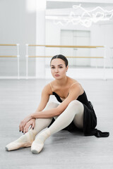 Fototapeta na wymiar brunette ballerina looking at camera while sitting on floor in studio
