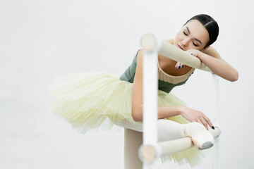 ballerina with closed eyes stretching leg on barre while exercising in studio