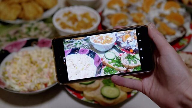 Female Hands Takes Photos, Videos of New Year's Table Food on a Smartphone. Home kitchen. There are a lot of cooked delicious salads, sandwiches, cake on the table. Blurred background. Close-up. 4K.