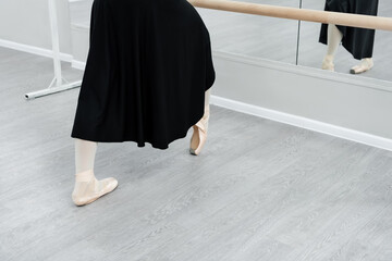 cropped view of ballerina in black dress training at barre in studio