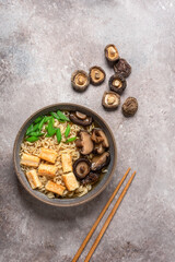 Asian vegan ramen noodle soup with roasted tofu cheese and shiitake mushrooms in a bowl on brown grunge background. Top view. Selective Focus.