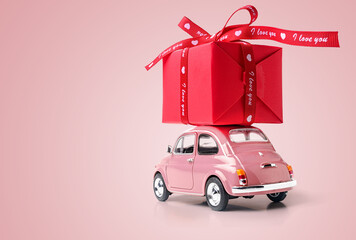 Pink retro toy car delivering red gift box with ribbon on pink background. Copy space.