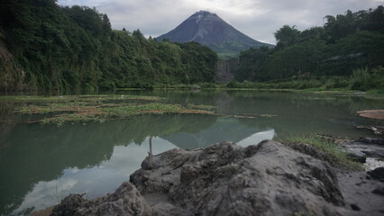 Lake with bluish water surrounded by trees and volcano on the background. The surface of the lake is overgrown by aquatic plants. The volcano named Merapi Volcano with cloudy weather