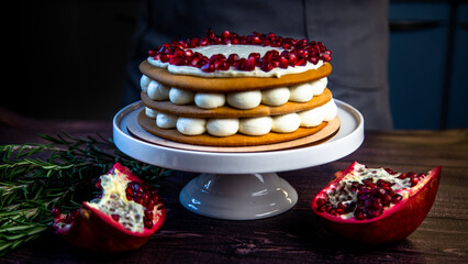 A cake of three layers, consisting of cakes and white cream, decorated with pomegranate and rosemary on top, stands on a white stand sideways on a dark background. High quality photo