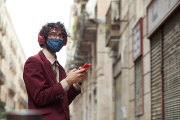 Happy millennial man smiling with mask after lock reopening - Hipster man with glasses enjoying with smart phone in hands and headphones on a Barcelona street. Copy-space
