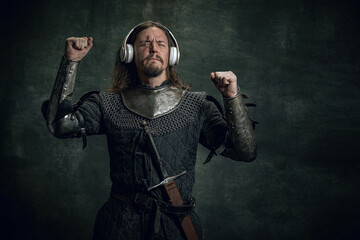 Young medieval warrior or knight with dirty wounded face in headphones listening to music isolated over dark vintage background. Comparison of eras, history