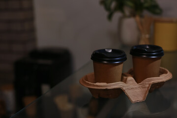Takeaway coffee cups with cardboard holder on glass table in cafe. Space for text