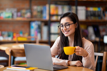 Happy latin woman working online at cafeteria, using laptop computer and drinking coffee, copy space