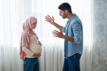 Angry arab man shouting at his pregnant muslim wife, standing near windiw at home, side view - obrazy, fototapety, plakaty