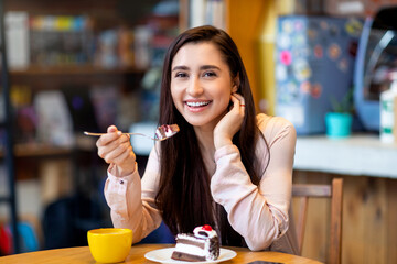 Delicious dessert. Happy arab lady eating cake and drinking coffee while resting in cafe and smiling at camera