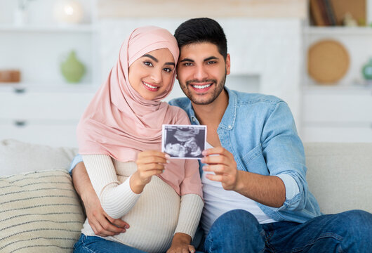 Happy pregnant muslim couple demonstrating baby sonography photo and smiling to camera