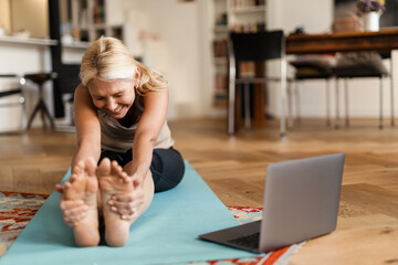 Blonde mature woman smiling and using laptop during yoga practice