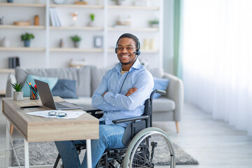 Positive paraplegic black man in wheelchair wearing headset, posing in front of laptop computer at...