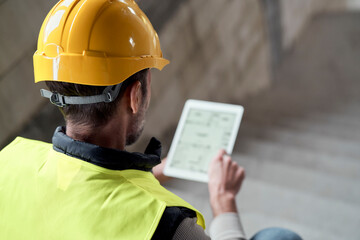 Caucasian engineer sitting on stairs and browsing digital tablet on construction site