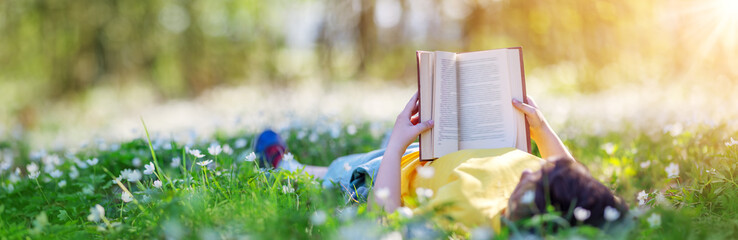 Small child lying down on the field in the spring park and reading a book - obrazy, fototapety, plakaty