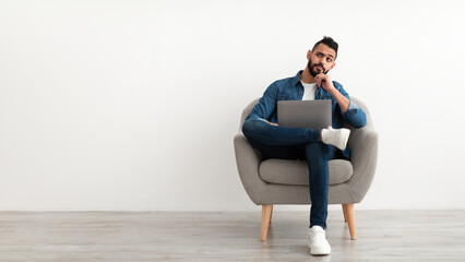 Thoughtful young Arab guy using laptop, sitting in armchair, looking aside at empty space against white studio wall