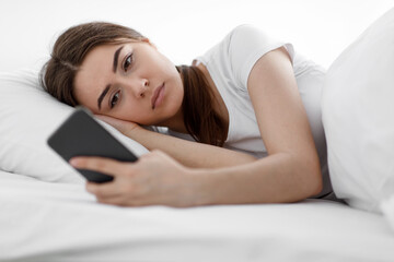 Obraz na płótnie Canvas Sad sorrowful caucasian millennial lady, reading message on smartphone, waiting for sms on bed