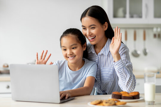 Happy asian mother and daughter having video call, waving and smiling at laptop screen, sitting in kitchen interior
